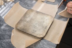 Base of a square baking tin on top of brown parchment paper which is being cut out with scissors to the shape of the tin